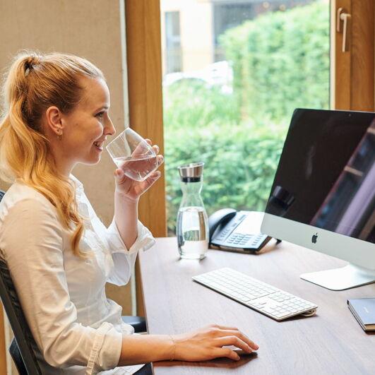 Frau sitzt am Schreibtisch mit einem Glas Wasser in der Hand