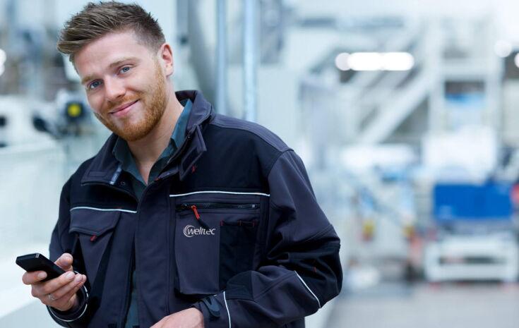 Junger Mann steht in Fabrik mit einem Handy in der Hand
