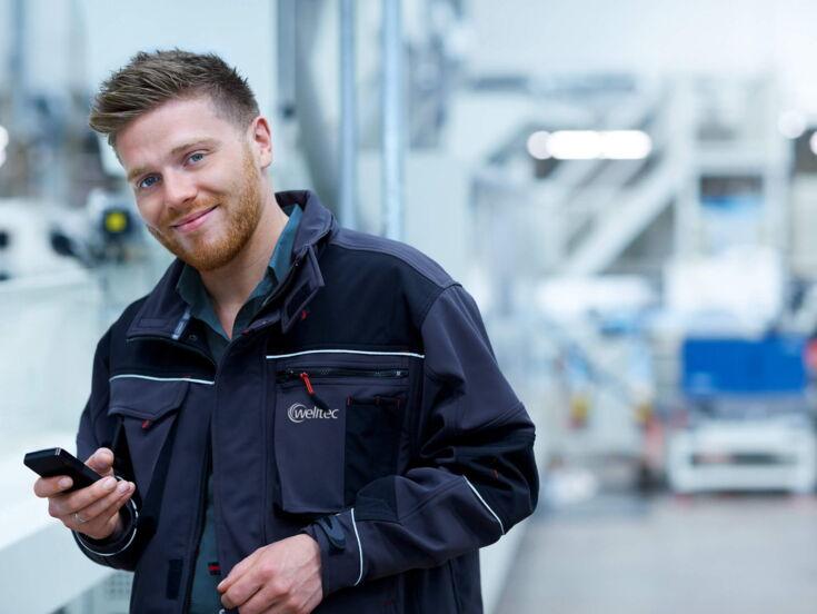 Junger Mann steht in Fabrik mit einem Handy in der Hand