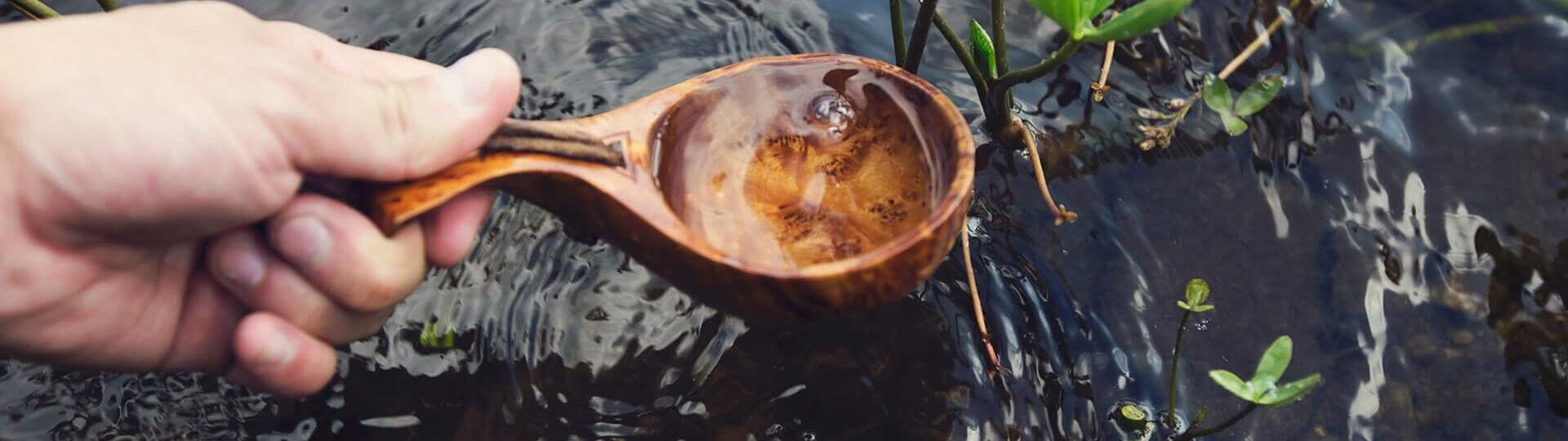 Wasser wird mit einem Holzloeffel aus einem Bach geschoepft