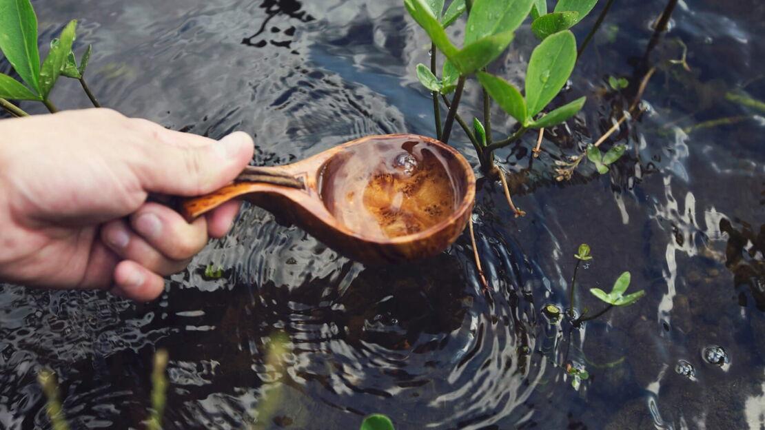 Wasser wird mit einem Holzloeffel aus einem Bach geschoepft
