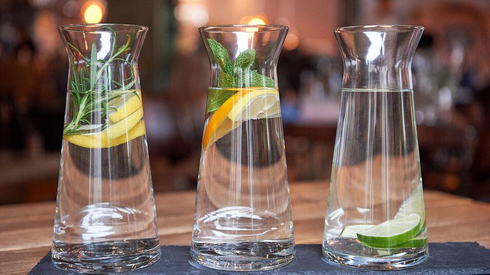 Drei Glas-Karaffen auf einem Schieferbrett, gefüllt mit Wasser und verschiedenen Zutaten: Rosmarin und Zitronenscheiben in der ersten Karaffe, Minzblätter und Zitronenscheiben in der zweiten Karaffe, Limettenscheiben in der dritten Karaffe. Die Szene spie